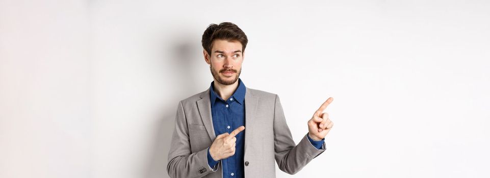 Doubtful male entrepreneur in suit looking and pointing left with hesitant face, standing unsure against white background.
