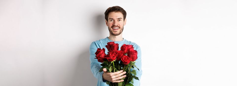 Handsome bearded guy stretch out hands, giving bouquet of roses and smiling, bring flowers on romantic date, celebrating Valentines day with lover, standing over white background.