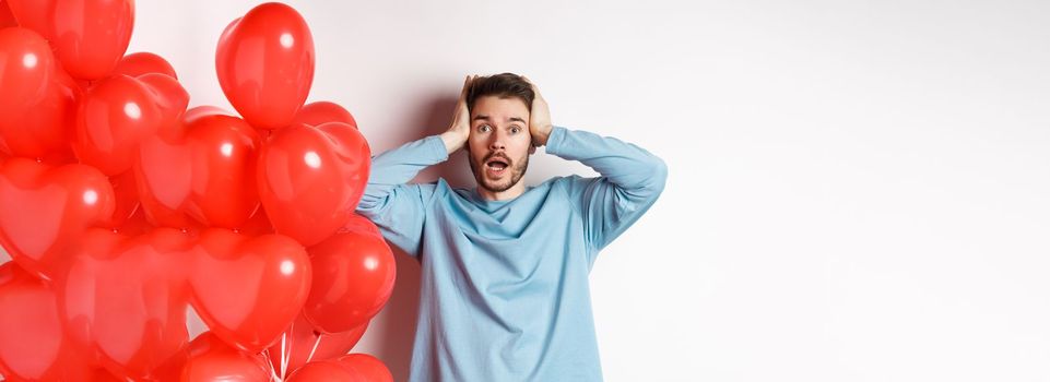 Confused boyfriend holding hands on head and panicking on Valentines holiday, alarmed with romantic presents on lovers day, standing near hearts balloon over white background.