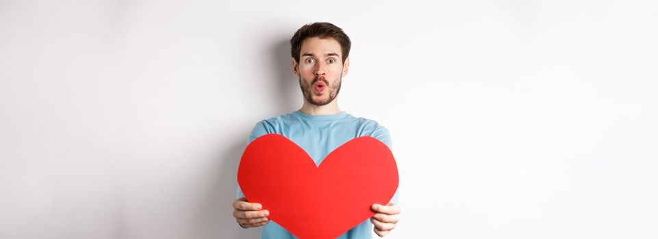 Young handsome man showing big red valentines day heart, being in love, pucker lips for kiss on valentine date, standing over white background.