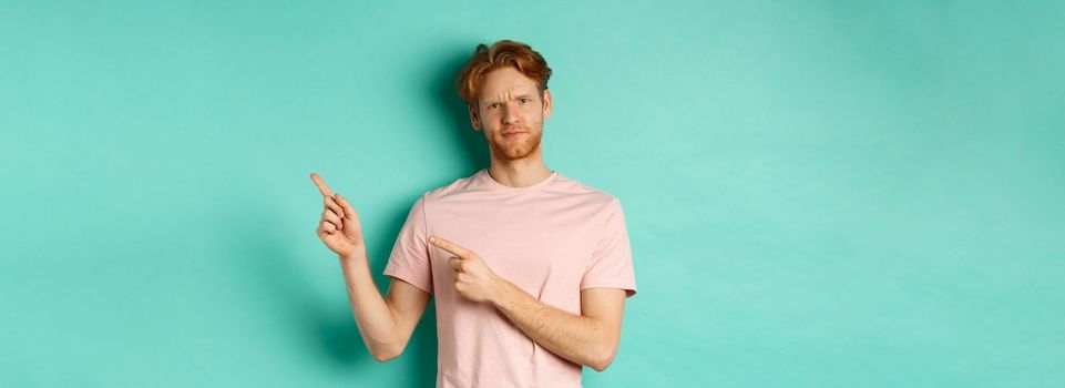 Skeptical and doubtful redhead man in t-shirt pointing fingers at upper right corner, showing promo offer with displeased face, standing over turquoise background.