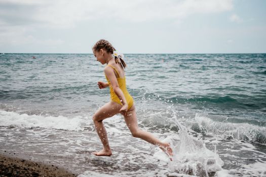 Cute little girl running along the seashore against a clear blue sea and rejoices in the rays of the summer sun. Beautiful girl in yellow swimsuit running and having fun on tropical beach