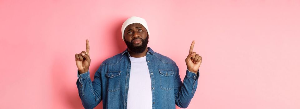 Upset and gloomy african american man in beanie, pointing fingers up, sighing and looking at top with sadness, standing over pink background.