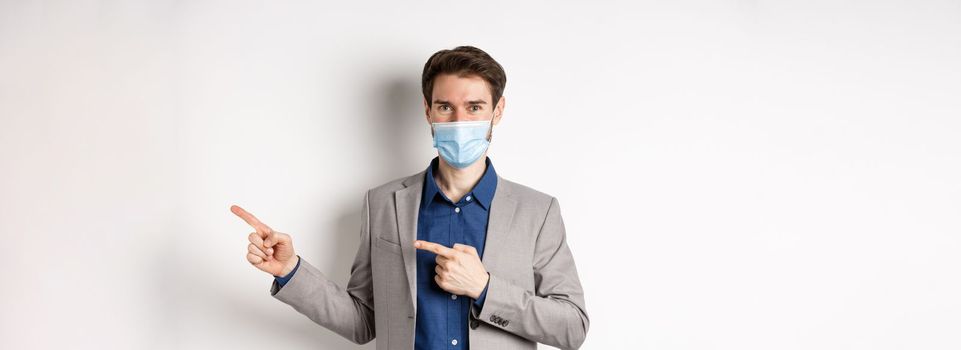 Covid-19, pandemic and business concept. Confident male manager in suit and medical mask showing way, pointing fingers right at advertisement, white background.