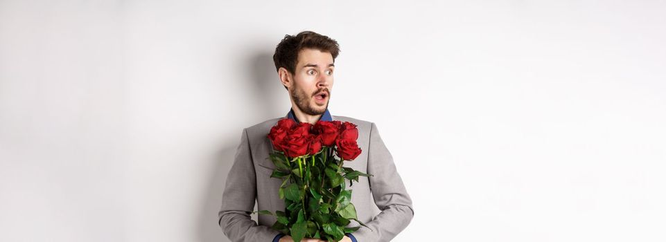 Handsome young man in suit holding red roses, looking left with surprised and startled expression, standing on Valentines day over white background.