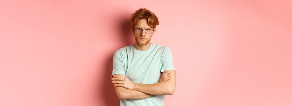 Angry man in glasses with red hair frowning, cross arms on chest in defensive pose, sulking at you, standing over pink background.