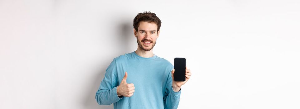 Smiling young man showing empty smartphone screen and thumbs up, praising good application, recommend mobile app, standing over white background.