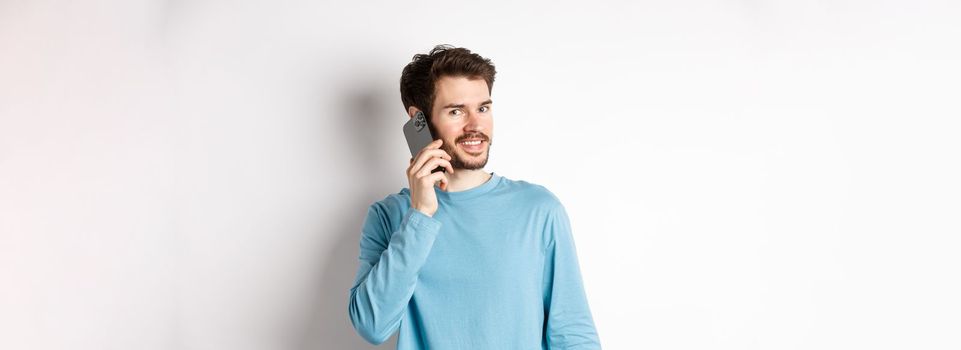 Technology concept. Young male model talking on mobile phone, calling someone on smartphone and smiling, standing over white background.