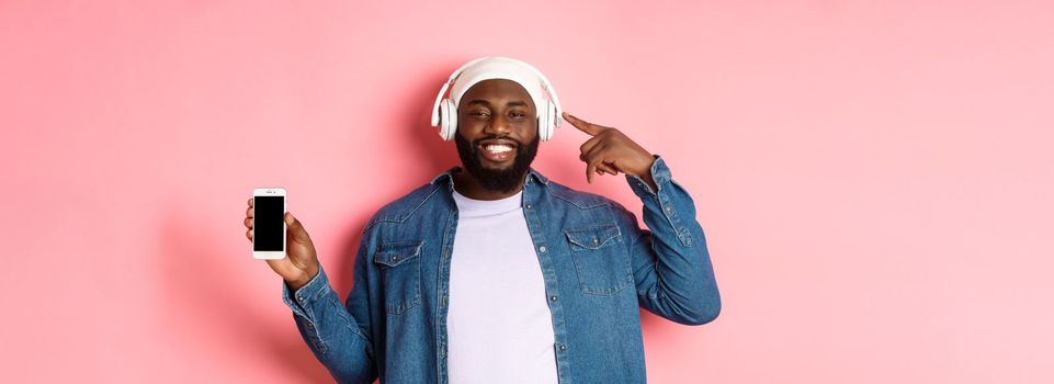 Happy black man listening music, pointing finger at headphones and smiling, showing mobile phone screen app or playlist, standing over pink background.