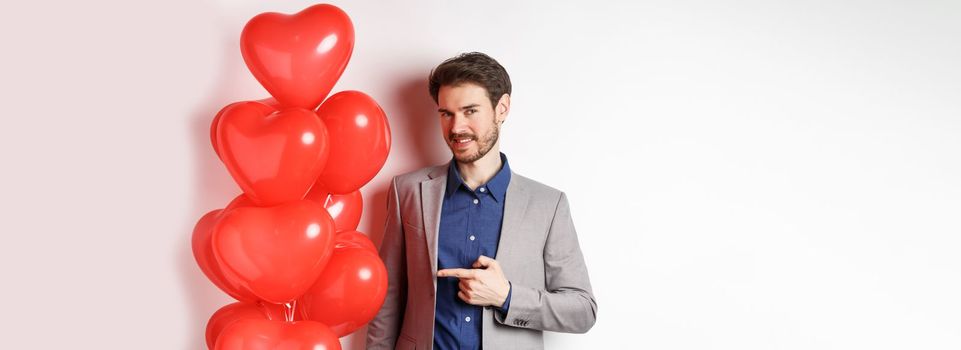 Lovers day. Charming young man with beard, wearing fancy suit, pointing finger at heart balloon surprise for Valentines day, standing over white background.