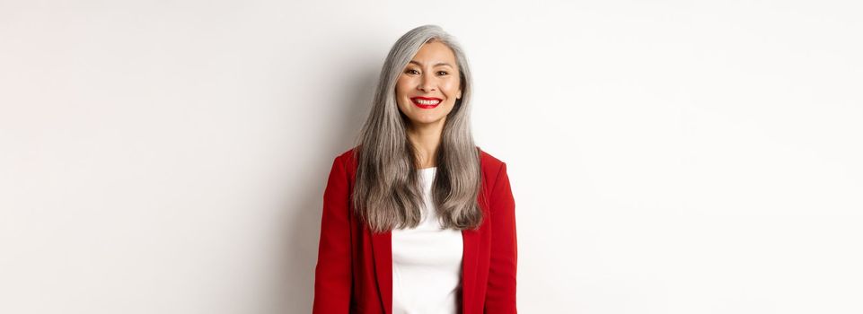 Business people. Senior smiling asian woman with red lipstick and blazer looking happy, standing over white background.