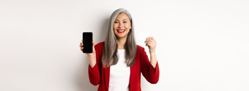 Senior asian businesswoman showing plastic credit card and blank smartphone screen, smiling at camera, white background.