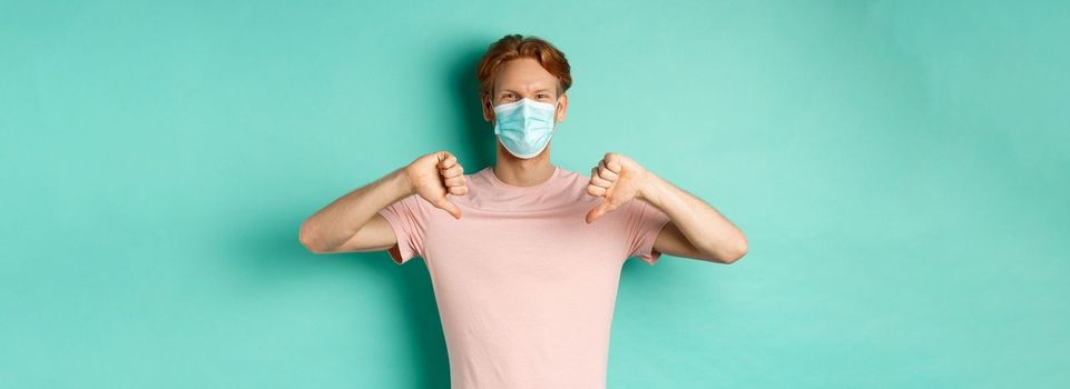 Covid-19, pandemic and lifestyle concept. Young man with red hair in face mask, showing thumbs down, dislike or disapprove something, standing over turquoise background.