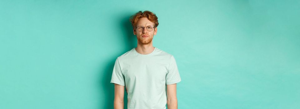 Confused young redhead man in glasses staring at camera puzzled, squinting perplexed, cant understand something, standing over mint background.
