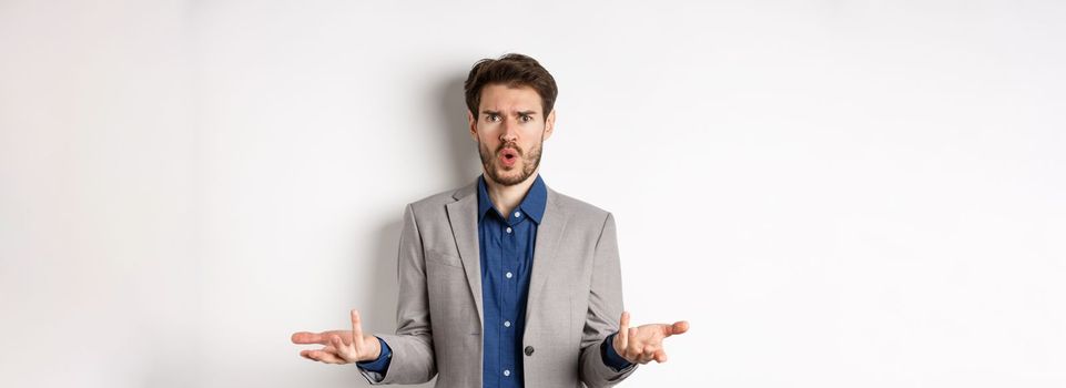 Confused and shocked businessman raising hands up and asking what happened, frowning disappointed, waiting for explanation, standing in suit on white background.