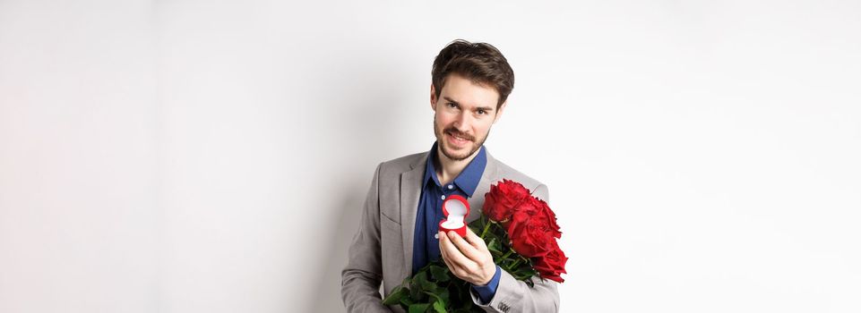 Romantic man with boquet of red roses asking to marry him, holding engagement ring and looking confident at camera, standing in suit over white background.