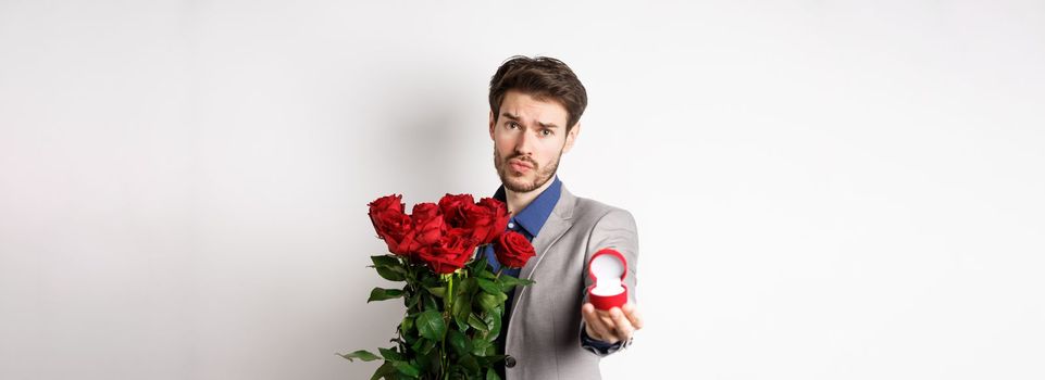 Handsome boyfriend in suit asking to marry him, standing with red bouquet of roses and engagement ring, looking pleading at camera, standing over white background.