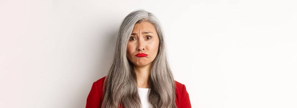 Close up of sad and unhappy asian senior woman sulking, frowning and looking jealous at camera, standing over white background.