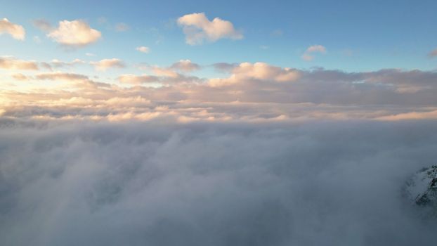 Flying among the ocean of clouds in the mountains. The fog is like big ocean waves crashing against high mountains covered with snow. Spruce trees grow in places. Sunset. Orange rays of the sun