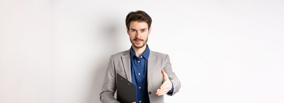 Confident businessman with clipboard giving hand for handshake, greeting business partners on meeting, standing on white background in suit.