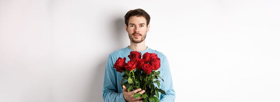 Confident young man bring flowers on Valentines day date, holding romantic bouquet, standing over white background.