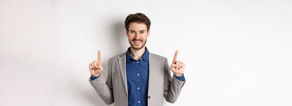 Confident successful businessman pointing fingers up, smiling and showing company logo, standing in suit on white background.