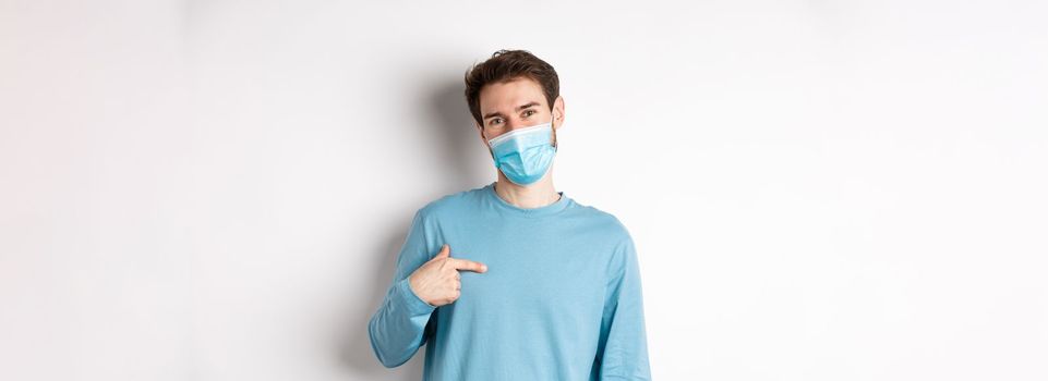 Covid-19, health and quarantine concept. Smiling caucasian man in medical mask pointing at himself, volunteering, standing over white background.