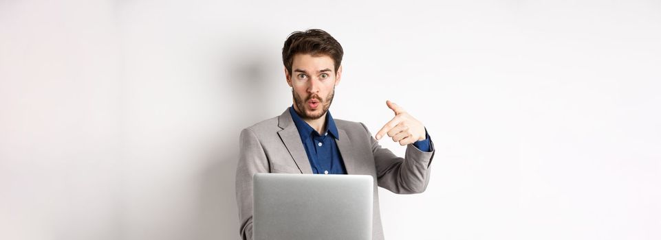Excited handsome man asking to look here, pointing at laptop screen with amazed face, standing in business suit against white background.