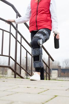 Woman wearing sport clothes and knee brace or orthosis after leg surgery, walking in the park. Medical and healthcare concept.