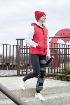 Woman wearing sport clothes and knee brace or orthosis after leg surgery, walking down the stairs in the park. Medical and healthcare concept.