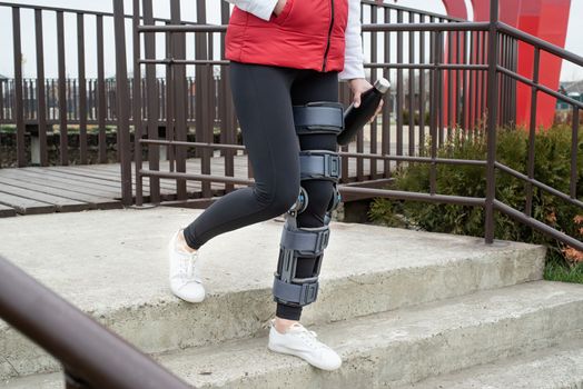 Woman wearing sport clothes and knee brace or orthosis after leg surgery, walking down the stairs in the park. Medical and healthcare concept.