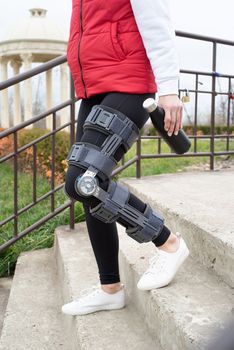 Woman wearing sport clothes and knee brace or orthosis after leg surgery, walking down the stairs in the park. Medical and healthcare concept.