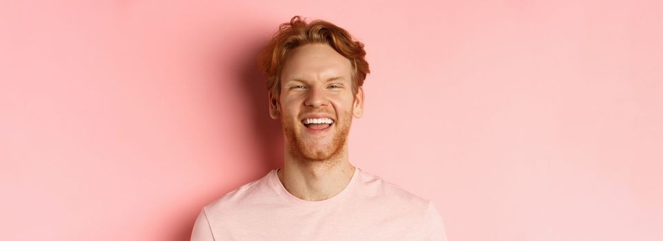 Close up of cheerful young man with messy red hair and beard, laughing and smiling, showing white teeth, looking happy at camera, standing over pink background.