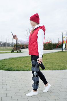 Woman wearing sport clothes and knee brace or orthosis after leg surgery walking in the park using smartphone. Medical and healthcare concept.