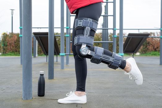 Woman wearing sport clothes and knee brace or orthosis after leg surgery, walking in the park. Medical and healthcare concept.