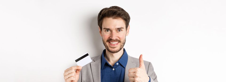 Shopping. Handsome satisfied client showing thumbs up and plastic credit card, smiling pleased, white background.