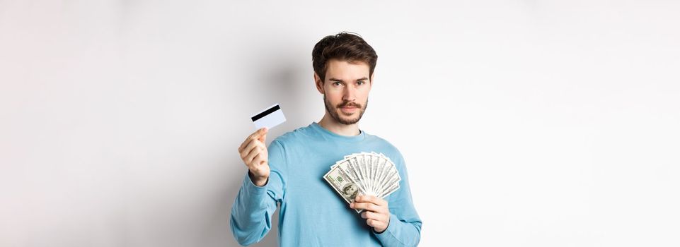 Caucasian man holding money and showing plastic credit card, standing in blue sweatshirt on white background.