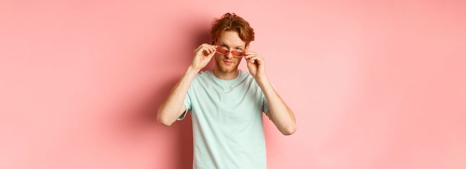 Handsome redhead man in summer sunglasses looking sassy at camera, standing over pink background.