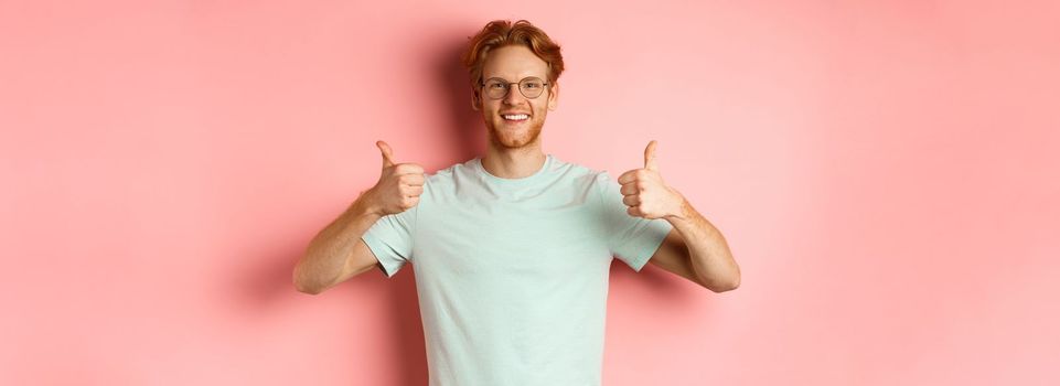 Cheerful european man with red hair and beard, wearing glasses, showing thumbs-up and smiling in approval, praise something good, standing over pink background.