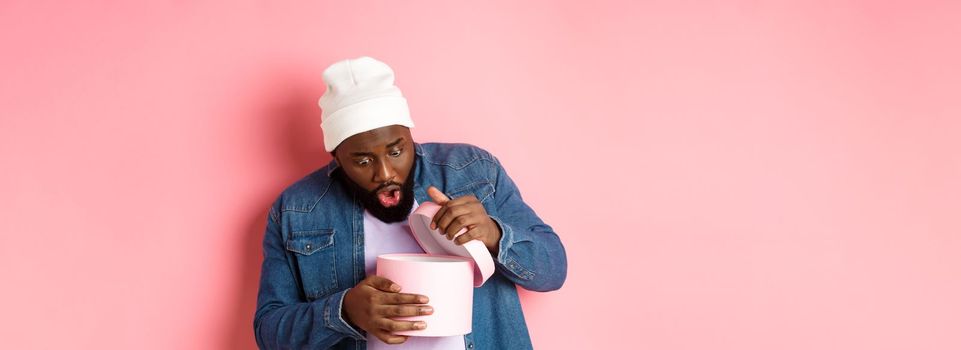 Image of surprised african-american man open box with birthday gift, staring at present with amazement, standing against pink background.