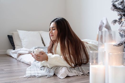 Merry Christmas and Happy New Year. Woman in warm white winter sweater lying in bed at home at christmas eve holding cup with marshmallows, fir tree behind