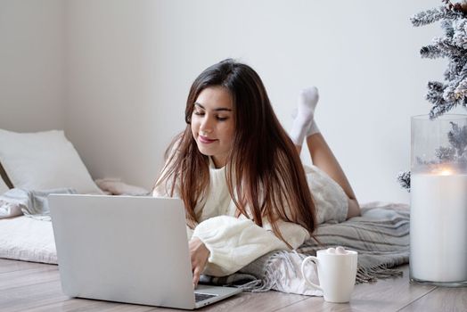 Merry Christmas and Happy New Year. Woman in warm white winter sweater lying in bed at home at christmas eve holding cup with marshmallows, fir tree behind