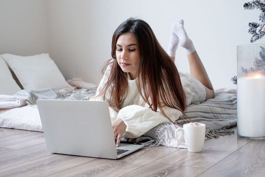 Merry Christmas and Happy New Year. Woman in warm white winter sweater lying in bed at home using laptop, watching movies or working