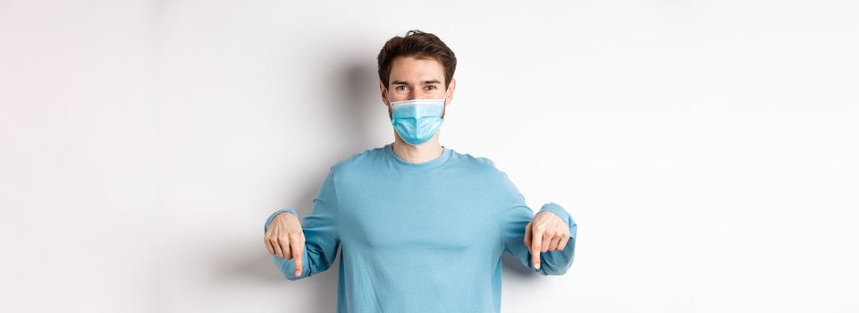 Covid-19, health and quarantine concept. Smiling caucasian man in face mask pointing fingers down, showing logo, standing over white background.