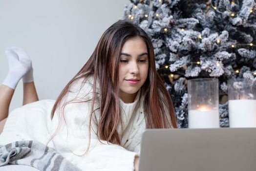 Merry Christmas and Happy New Year. Woman in warm white winter sweater lying in bed at home at christmas eve holding cup with marshmallows, fir tree behind