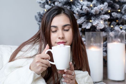 Merry Christmas and Happy New Year. Woman in warm white winter sweater lying in bed at home at christmas eve holding cup with marshmallows, fir tree behind