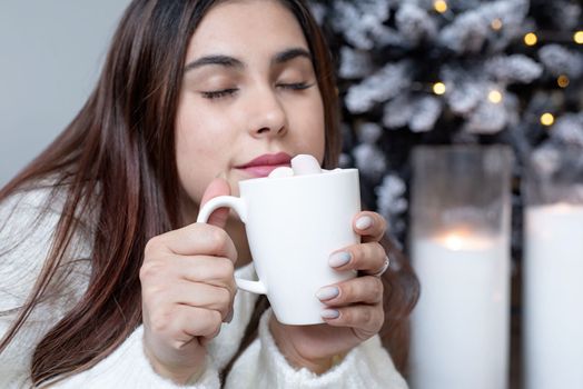 Merry Christmas and Happy New Year. Woman in warm white winter sweater lying in bed at home at christmas eve holding cup with marshmallows, fir tree behind