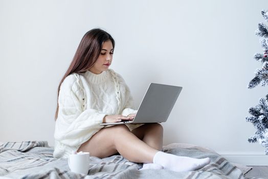 Merry Christmas and Happy New Year. Woman in warm white winter sweater lying in bed at home using laptop, watching movies or working