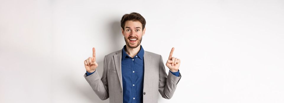 Excited lucky businessman in suit pointing fingers up, smiling amused, showing cool advertisement, standing on white background.