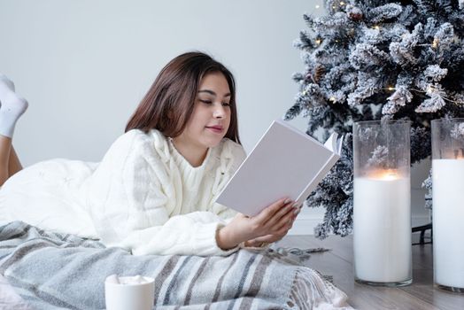 Merry Christmas and Happy New Year. Woman in warm white winter sweater lying in bed at home at christmas eve holding cup with marshmallows, fir tree behind